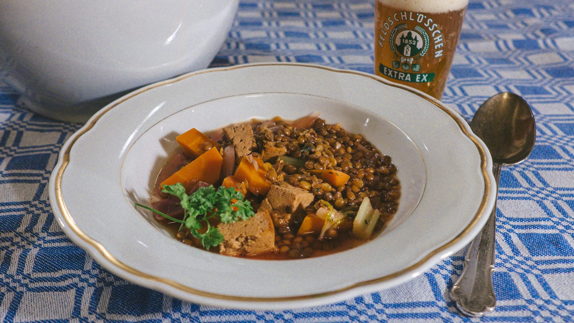 Ein Foto der Rote Beete, Möhren und Linsensuppe. Die Suppe steht angerichtet auf einem Teller mit Goldrand, daneben steht ein kleines vintage Bierglas der Marke Feldschlösschen aus Braunschweig.