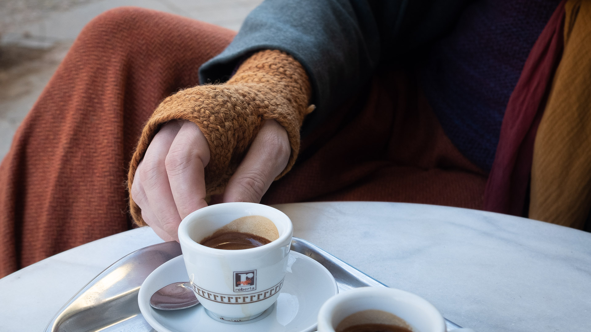 Merlins Hand verpackt in einer orange Stulpe greit nach einer Tasse Espresso